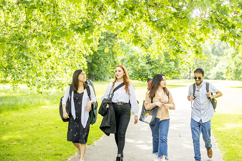 A group or students, holding bags and musical instruments, walking in a park, with green leaves on a tree.